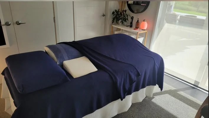 Lydia's treatment room: a massage table made up with navy covers, pillows and a white valance, beside a large window and a shelf with a softly glowing lamp